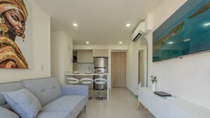 Uma sala de estar moderna com um sofá cinzento, cozinha com armários brancos e área de jantar com cadeiras brancas.