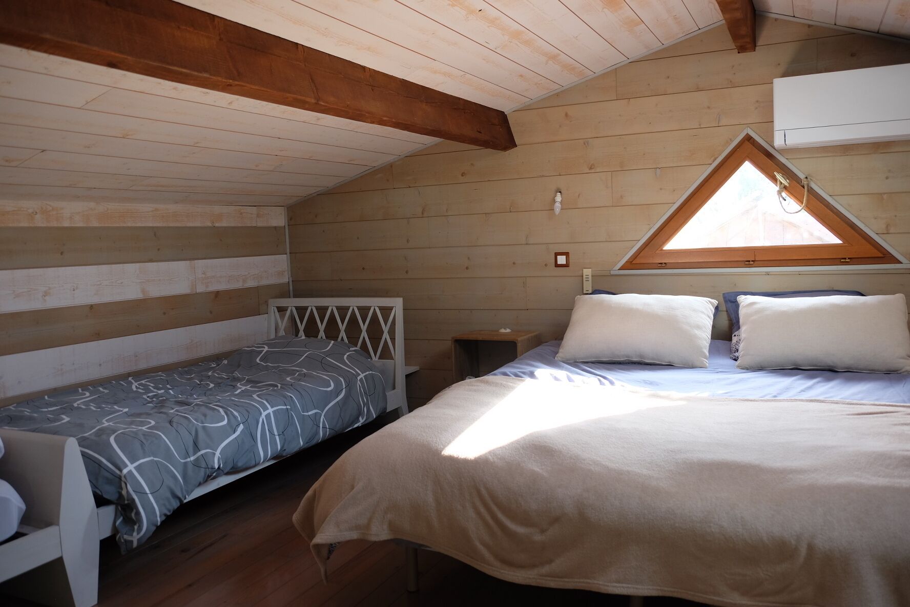 A small, cozy room with a sloped ceiling and wooden walls. There are two beds with white headboards and a small table. A window with a triangular wooden frame allows natural light into the room.