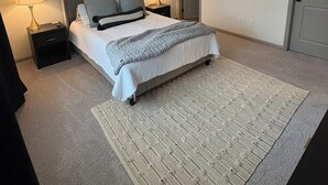 Un dormitorio con cama, dos mesitas de noche con lámparas, ventilador de techo, alfombra y cuadros en la pared.
