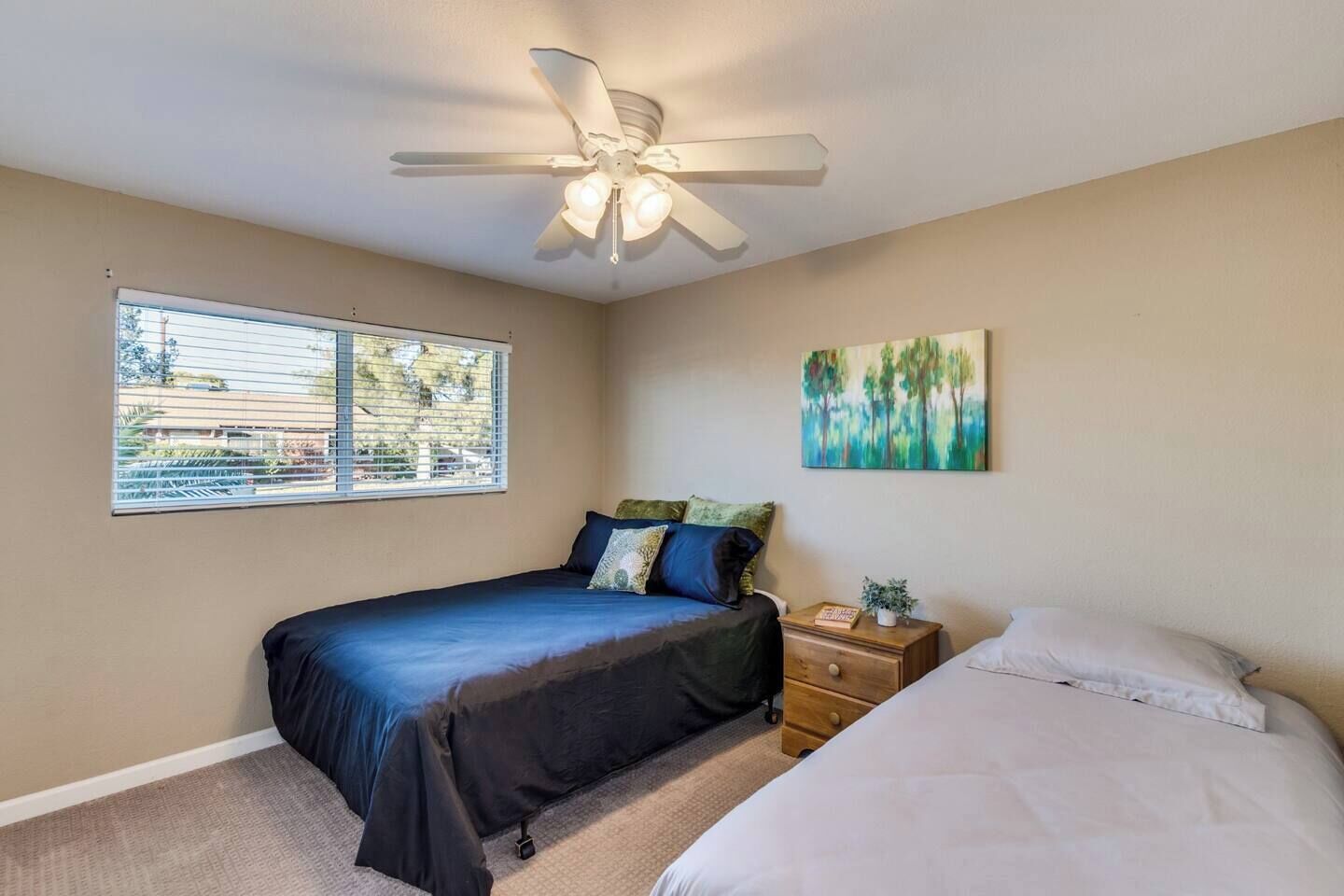 A bedroom with a bed, a nightstand, a ceiling fan, a window, and a painting on the wall.