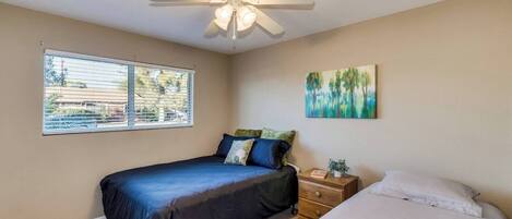 A bedroom with a bed, a nightstand, a ceiling fan, a window, and a painting on the wall.