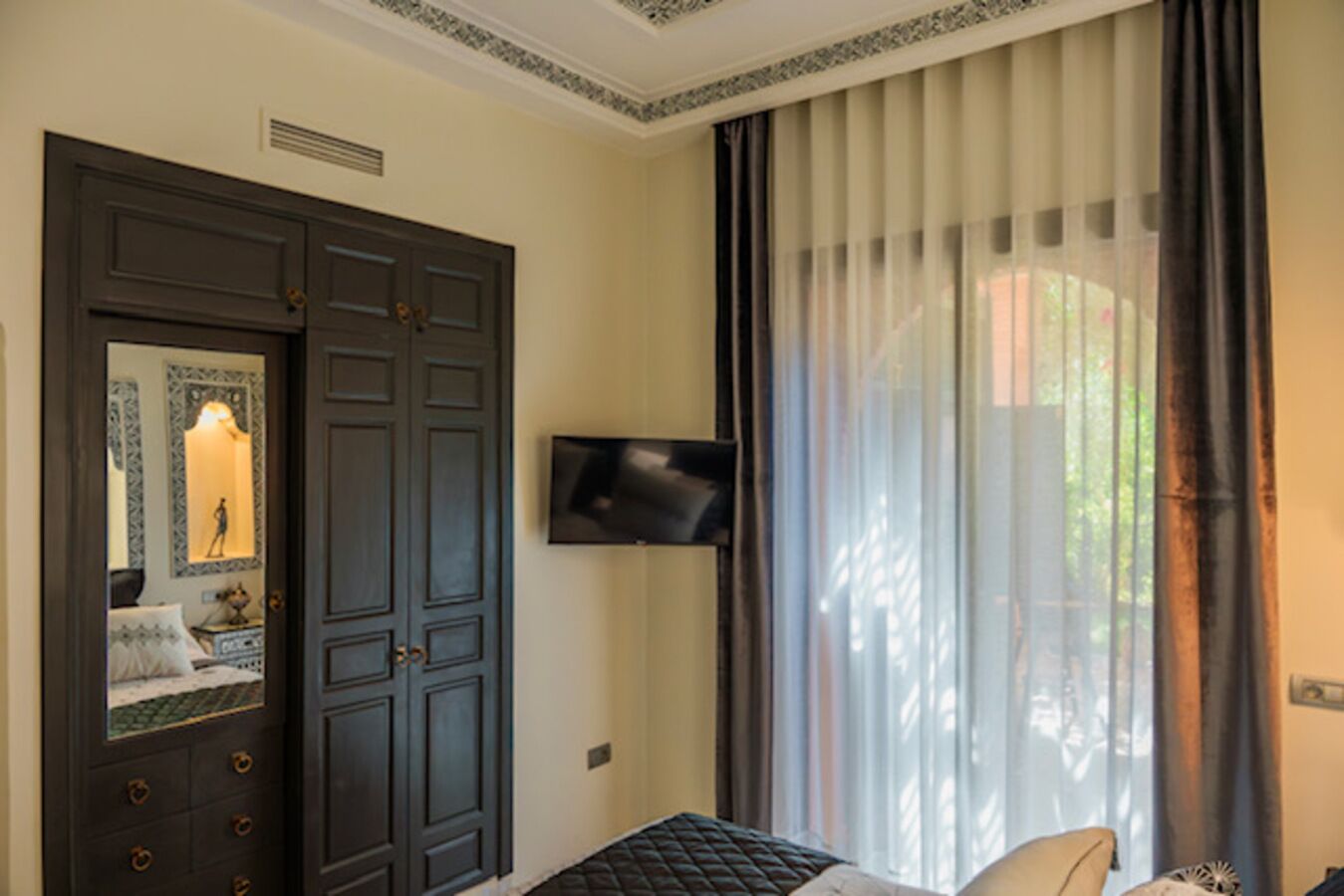 Habitació d'hotel amb una porta de fusta fosca gran, un televisor pantalla plana fixat a la paret i un finestral amb cortines primes.