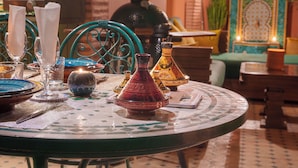 Ein runder Tisch mit einem Tischläufer aus Mosaik, umgeben von marokkanischen Dekorationselementen wie Teekanne, Kerze und Vase.