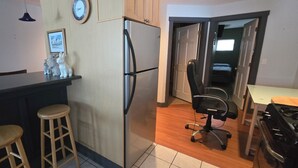 A kitchen with a refrigerator, a wooden counter, two stools, a chair, and a clock on the wall.