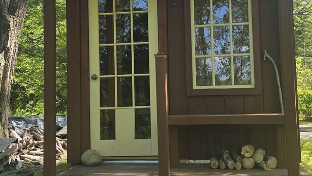 A small wooden cabin with a front door, a window, and a porch with logs.