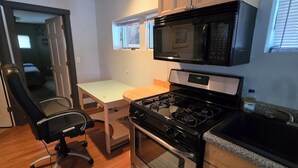 A compact kitchen with a stove, microwave, and sink. There's a chair and a small table.