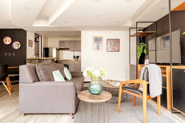 Een moderne woonkamer met een grijze bank, een houten salontafel en een klein bijzettafeltje. Achtergrond is een keuken met houten kastjes en een roestvrijstalen koelkast.