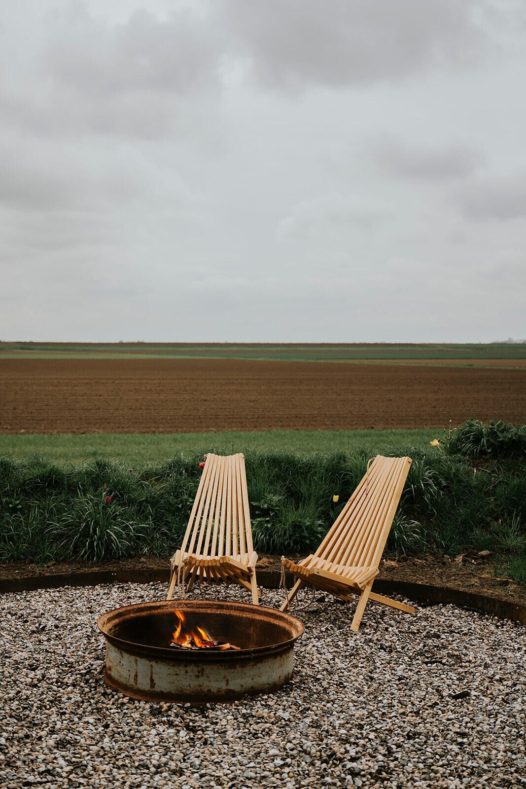 Two wooden lounge chairs facing a fire pit with a view of a field.