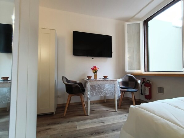 A small, modern room with a wooden floor, a white bed, a table with a vase of flowers, two chairs, a flat-screen TV mounted on the wall, and a window with a fire extinguisher below it. - Lovely newly renovated studio apartment in strategic location (Milano)
