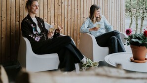 Twee personen zitten op witte stoelen in een kamer met houten paneelwanden, een ronde tafel en een potplant.
