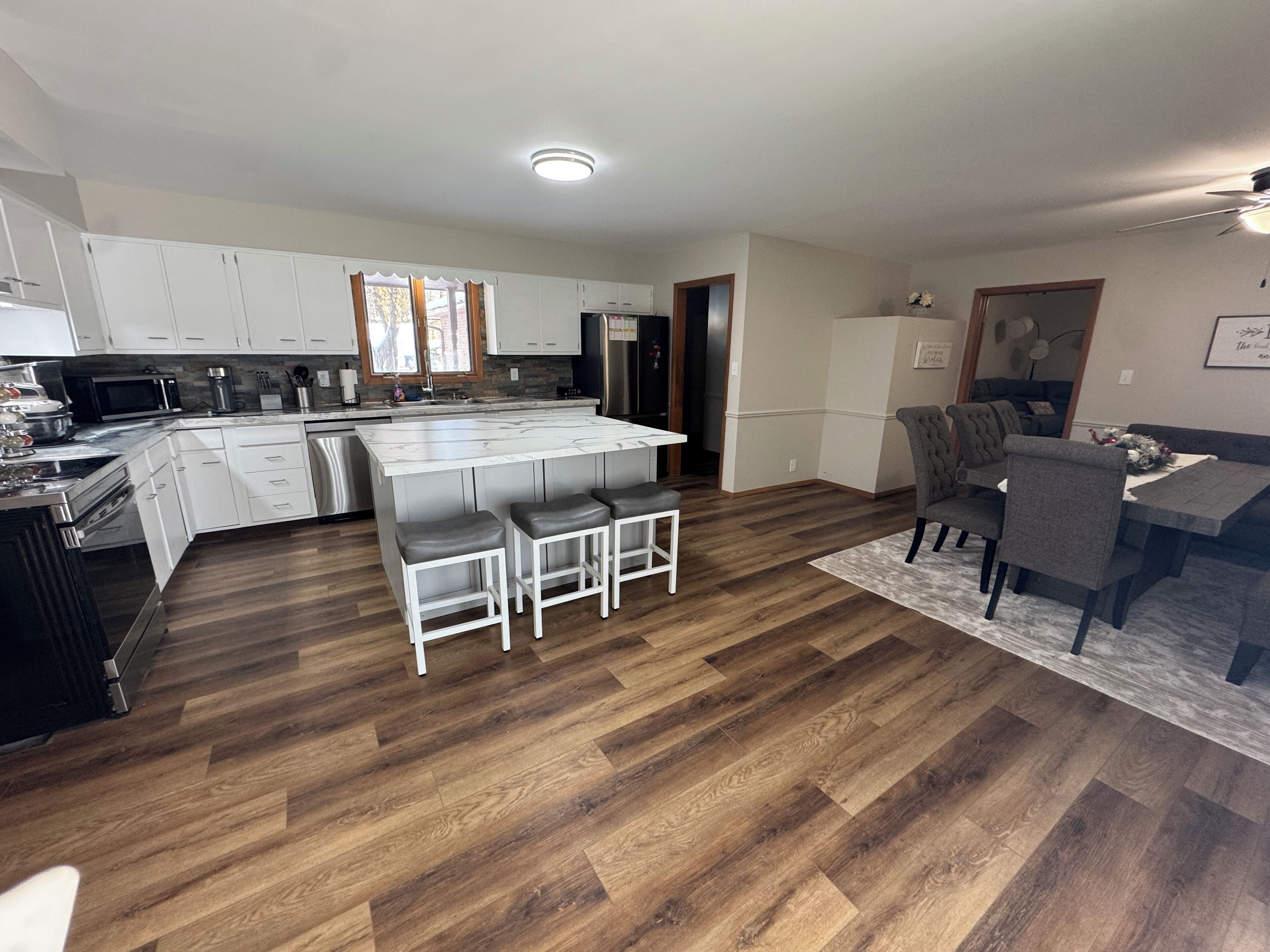 Une cuisine moderne avec des meubles blancs, un îlot central doté de tabourets, un coin repas avec une table et des chaises grises, et un parquet en bois disposé en motif chevron.