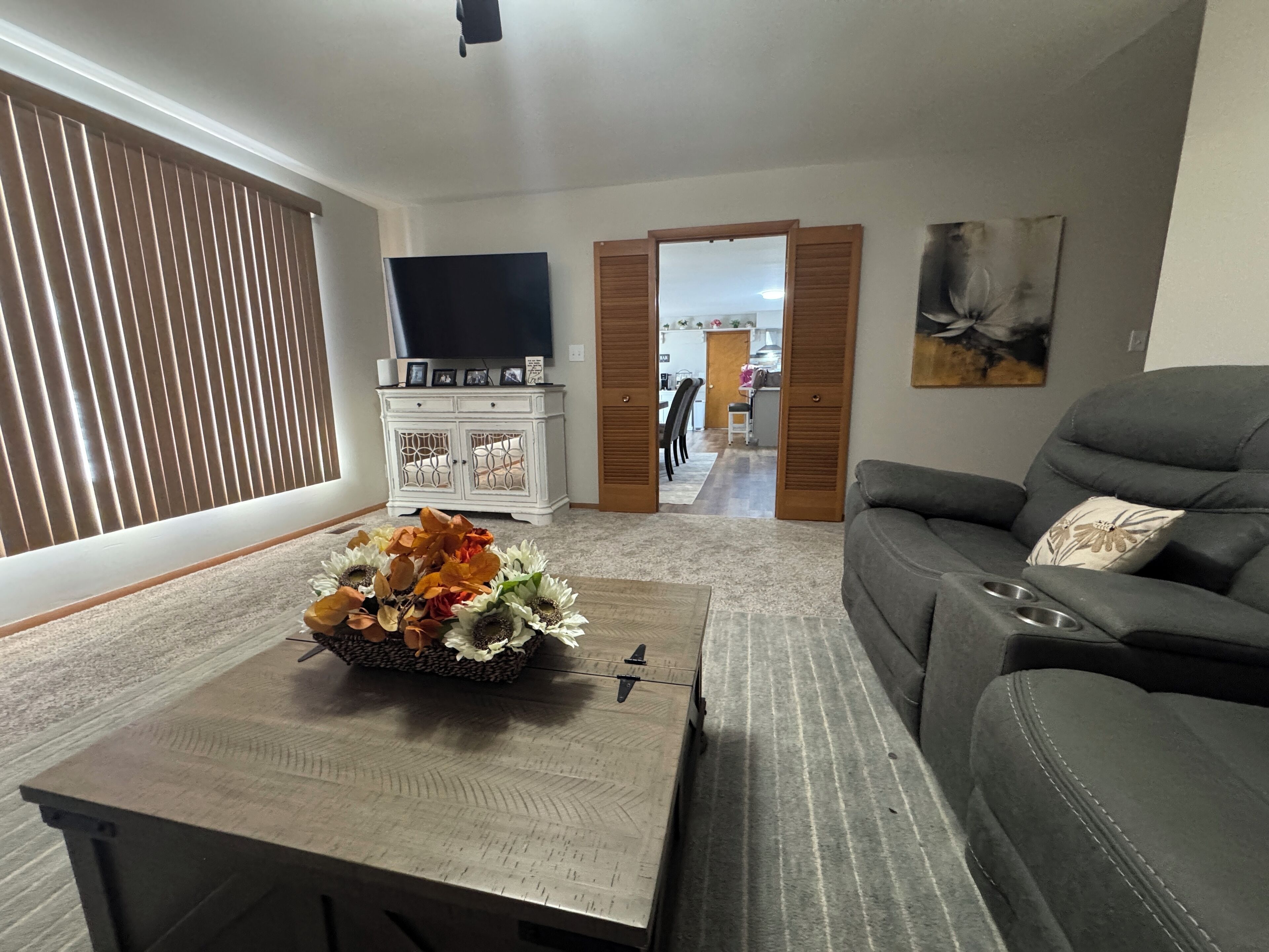 Un salon avec un canapé, une table basse agrémentée d’un arrangement floral, un téléviseur fixé au mur et une porte donnant sur une autre pièce.