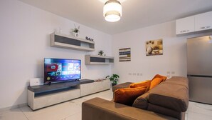 Ett modernt vardagsrum med en platt-TV, en soffa, ett soffbord och ett inbyggt köksområde med ett kylskåp.
