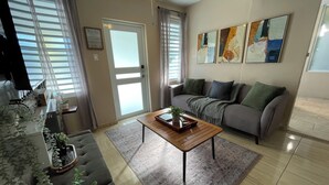 Ein Wohnzimmer mit grauem Sofa, einem Holztisch und einem grauen Teppich. Auf dem Sofa stehen Blumentöpfe und an der Wand hängt ein Gemälde.