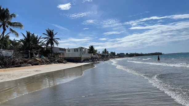 Ein Strand mit blauem Himmel, ruhiger See und einigen Gebäuden am Ufer.
