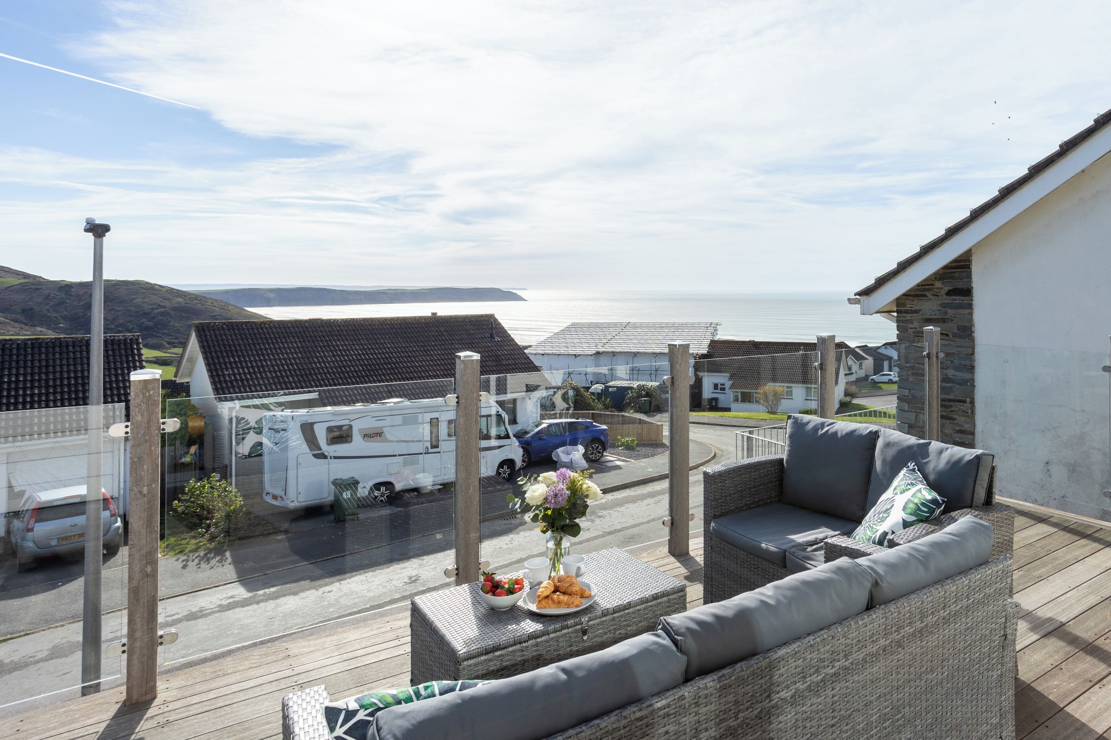 A balcony with wicker furniture, a dining table with food, and a view of the sea and parked vehicles.