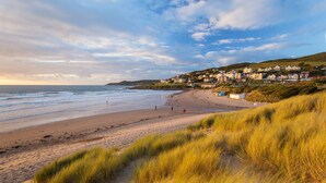 A beach with a grassy foreground, a sandy shore, and houses in the background. - Stylish coastal home with stunning sea views (Woolacombe)