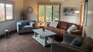 A cozy living room with a high ceiling, a fireplace, a large window with a view of trees, and a comfortable seating area with a coffee table.