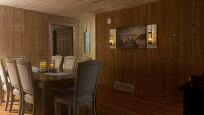 A wooden-paneled dining room with a wooden table and chairs, a painting on the wall, and two wall-mounted lanterns.