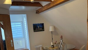 Un comedor con una mesa redonda de vidrio, cuatro sillas, una mesita auxiliar con una lámpara y un cuadro en la pared.