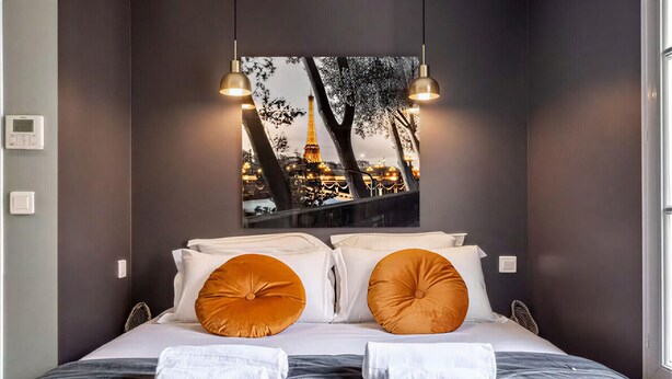 Ein Hotelzimmer mit einem Bett, zwei Kissen, einer Decke und einem an der Wand hängenden Bild des Eiffelturms.
