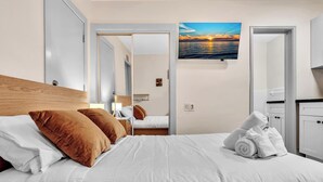 Ein ordentlich bezogenes Bett mit weißen Leinen und braunen Kissen, ein Nachttisch mit Lampe und Blick auf das Meer durch ein großes Fenster.