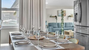 A dining table set with plates and wine glasses, a modern kitchen with stainless steel appliances, and a living area with a blue sofa and decorative plants. - Santorini Blu | Spacious 2BR Beachfront Suite (Hollywood)