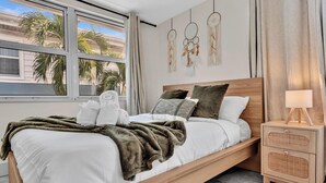 A modern bedroom with a bed, bedside table, and a view of palm trees through large windows. - Santorini Blu | Spacious 2BR Beachfront Suite (Hollywood)