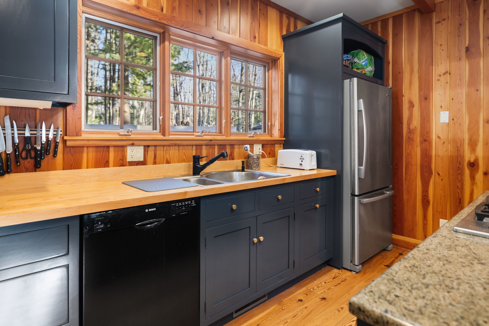 Una cocina con paredes de madera, un refrigerador de acero inoxidable, un lavavajillas Bosch y un ventanal con vistas a árboles.