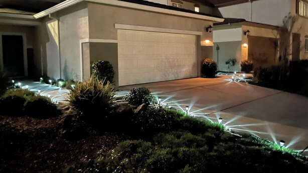 A residential driveway with a garage, surrounded by landscaped greenery and illuminated by ground lights.