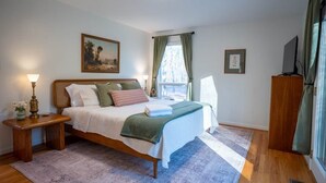 Een slaapkamer met een houten bed, een nachtkastje met een lamp, een raam met gordijnen, en een schilderij aan de muur.