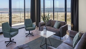 A spacious living room with a large window overlooking a beach, furnished with a sofa, armchair, ottoman, and a coffee table. - Apartment in Breskens Near the Beach (Breskens)