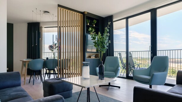 A modern living room with a glass table, blue chairs, a sofa, and a balcony with a view. - Apartment in Breskens Near the Beach (Breskens)