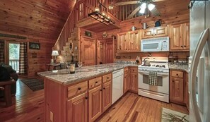 A wooden cabin interior with a kitchen featuring wooden cabinets, a granite countertop, a stove, a microwave, and a refrigerator. - Creekside Ellijay Cabin,  Hot Tub, Trout Stream, Outdoor Fireplace (Ellijay)