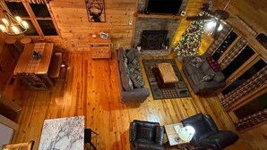 A cozy log cabin interior with a wooden floor, a stone fireplace, a Christmas tree, and a dining area with a table and chairs.