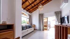 Ein modernes Wohnzimmer mit Holzboden, einer Couch mit gemusterten Kissen, einem Fernseher auf einem Holzschrank und einem großen Fenster mit Vorhängen.