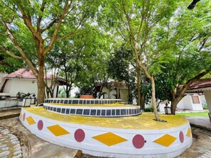 A circular outdoor stage with a yellow and white color scheme, surrounded by trees and a building with a red-tiled roof. - Malgudi Days and Resorts (Ujjain)