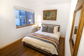 Un dormitorio con cama, ventana, cuadro y mesita de noche.