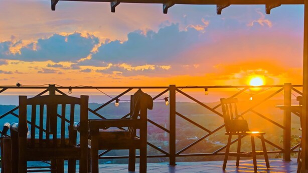 Une terrasse en bois, dotée de chaises et d’une rambarde, qui offre une vue sur un paysage au coucher du soleil.