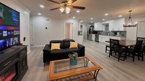 A modern living room with a flat-screen TV, a ceiling fan, a black sofa, a glass coffee table, and a dining area with a table and chairs. - Near AT&T • FIFA & Rangers • Sleeps 8 • Modern Home • Big Yard (Arlington)