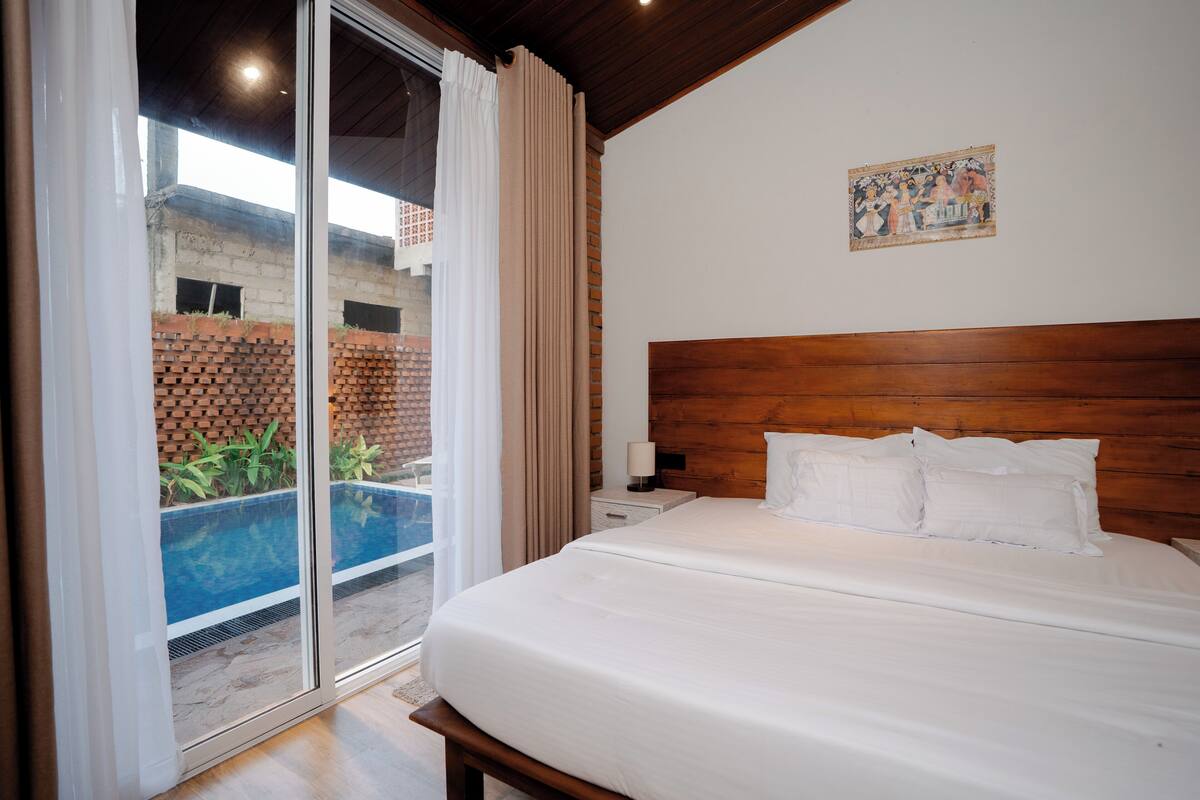 A hotel room with a large bed, a wooden headboard, a nightstand, a lamp, and a view of a swimming pool through a glass door.