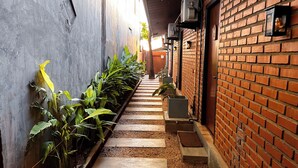 Un couloir étroit, avec un mur de briques d’un côté et un mur de béton de l’autre, bordé de plantes en pot et d’un chemin en bois.