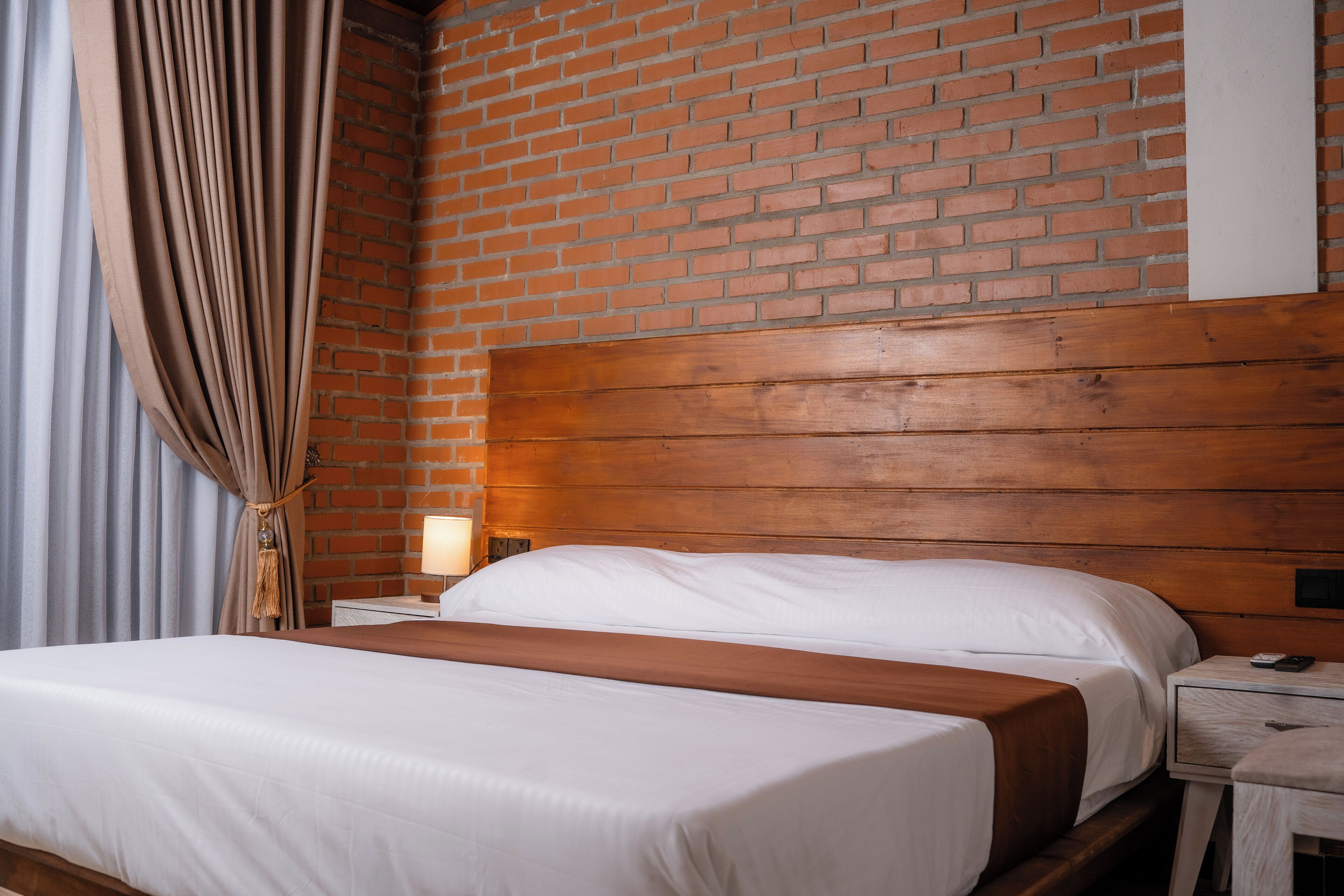 A hotel room with a brick wall, wooden headboard, two beds with white linens, a nightstand with a lamp, and a curtain.