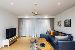 A modern living room with a flat-screen TV, a dark-colored sofa with colorful cushions, a wooden coffee table, and a ceiling fan. - Suite Azure 2br | Laguna beachside bang tao beach (Choeng Thale)
