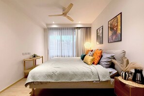 A bedroom with a bed, a bedside table, a ceiling fan, a window with curtains, and wall art. - Suite Azure 2br | Laguna beachside bang tao beach (Choeng Thale)