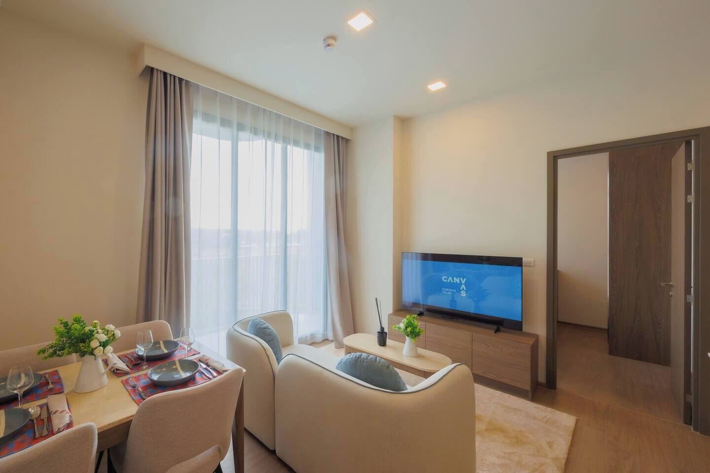 A modern living room with a flat-screen TV, a dining area with a table set for two, and a large window with curtains.