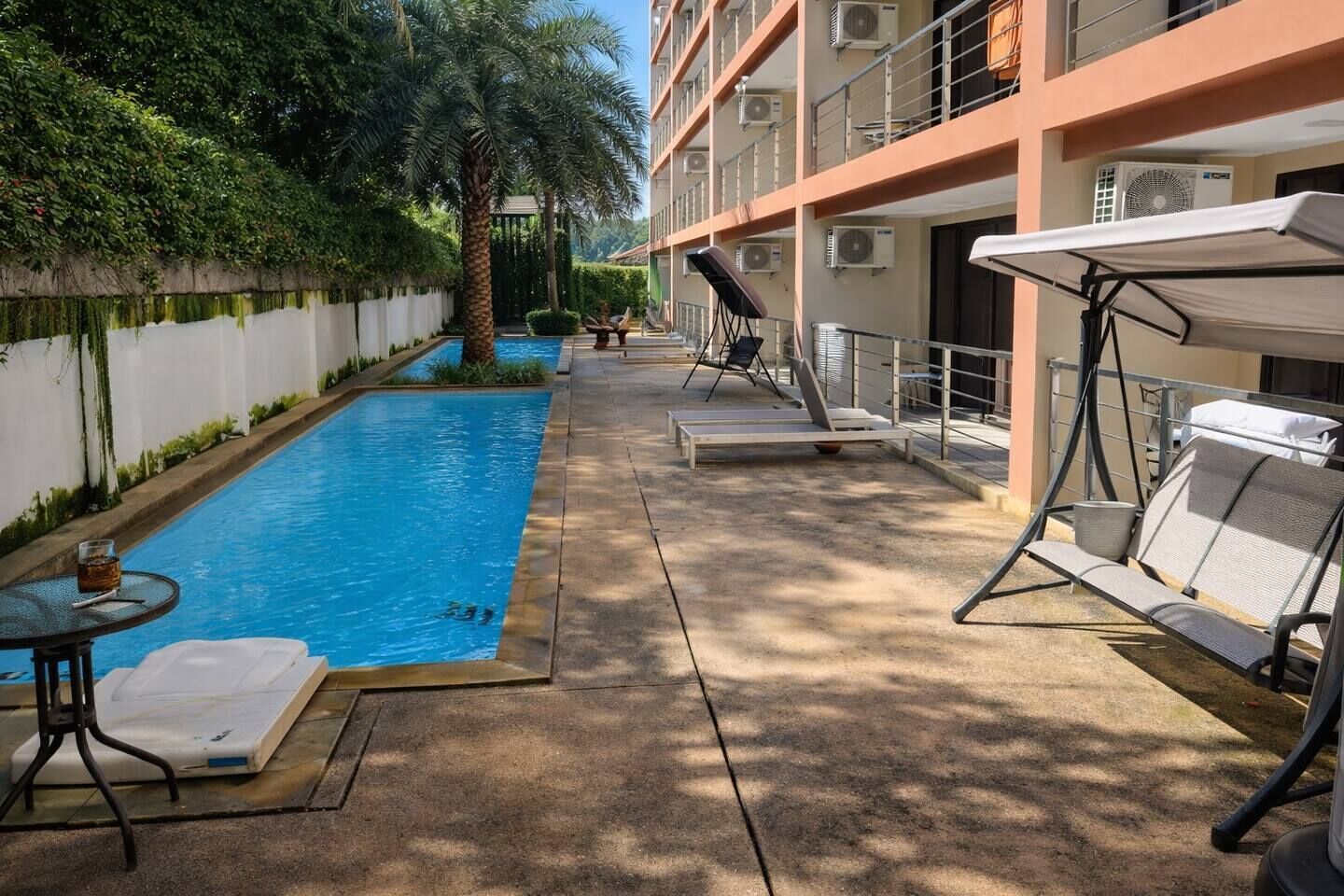 A pool area with lounge chairs and a table, surrounded by a building with balconies and air conditioning units.