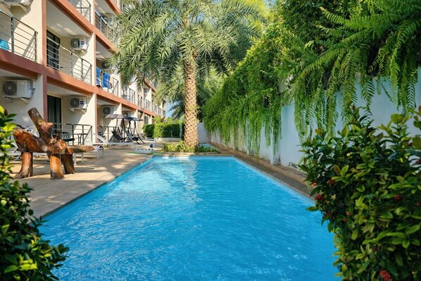 A swimming pool with a palm tree and greenery, surrounded by a building with balconies and outdoor furniture.