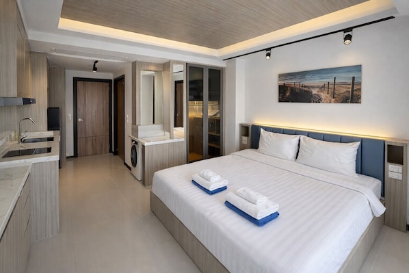 A modern hotel room with a large bed, a built-in washing machine, a vanity with a mirror, and a painting on the wall.