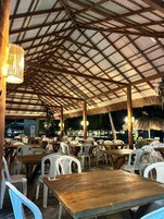 Un restaurante al aire libre con mesas de madera y sillas de plástico blanco, cubierto por un techo de metal con una sección de techo de paja.